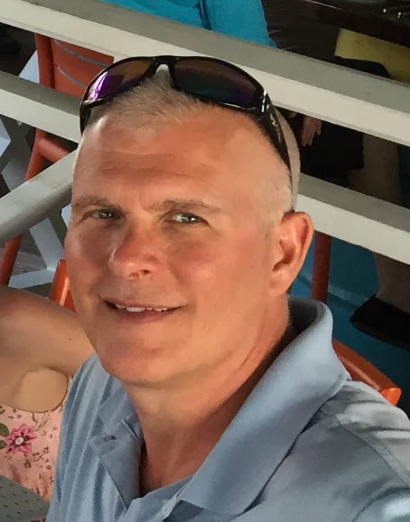 A smiling middle-aged man with short gray hair wearing a blue polo shirt and sunglasses.